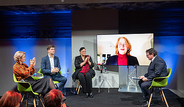 Das Bild zeigt die Podiumsdiskussion, welche im Rahmen der Veranstaltung umgesetzt wurde. Auf dem Podium fünf Personen, von denen eine über einen Bildschirm zugeschaltet ist.