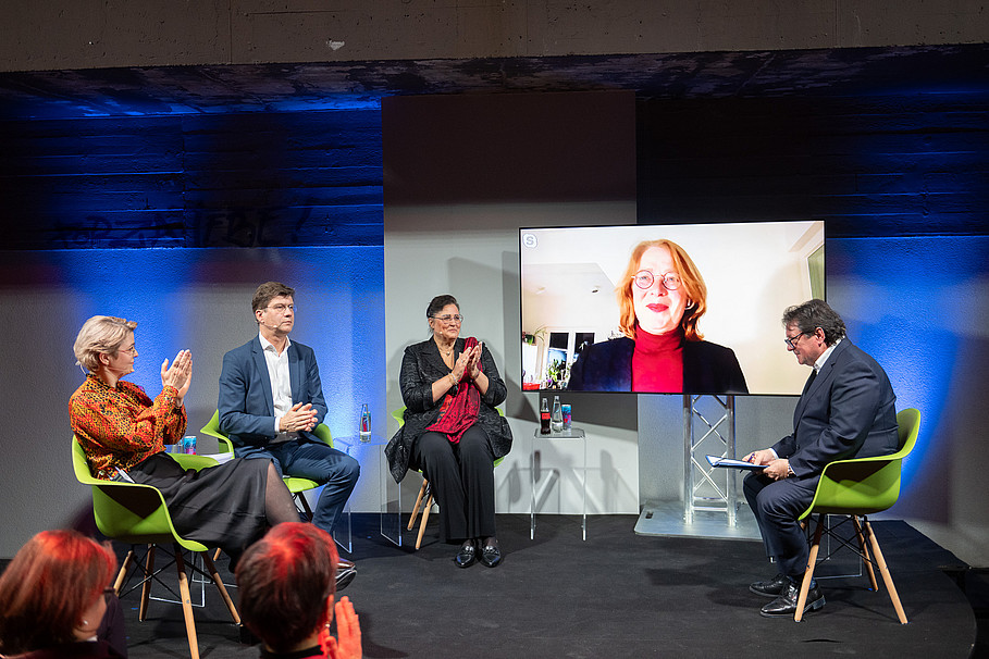 Das Bild zeigt die Podiumsdiskussion, welche im Rahmen der Veranstaltung umgesetzt wurde. Auf dem Podium fünf Personen, von denen eine über einen Bildschirm zugeschaltet ist.