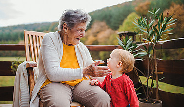 Eine ältere Frau sitzt auf einer Holzveranda und lächelt ein kleines Kind an, das vor ihr steht. Beide wirken fröhlich und liebevoll verbunden, im Hintergrund sind herbstliche Bäume zu sehen.