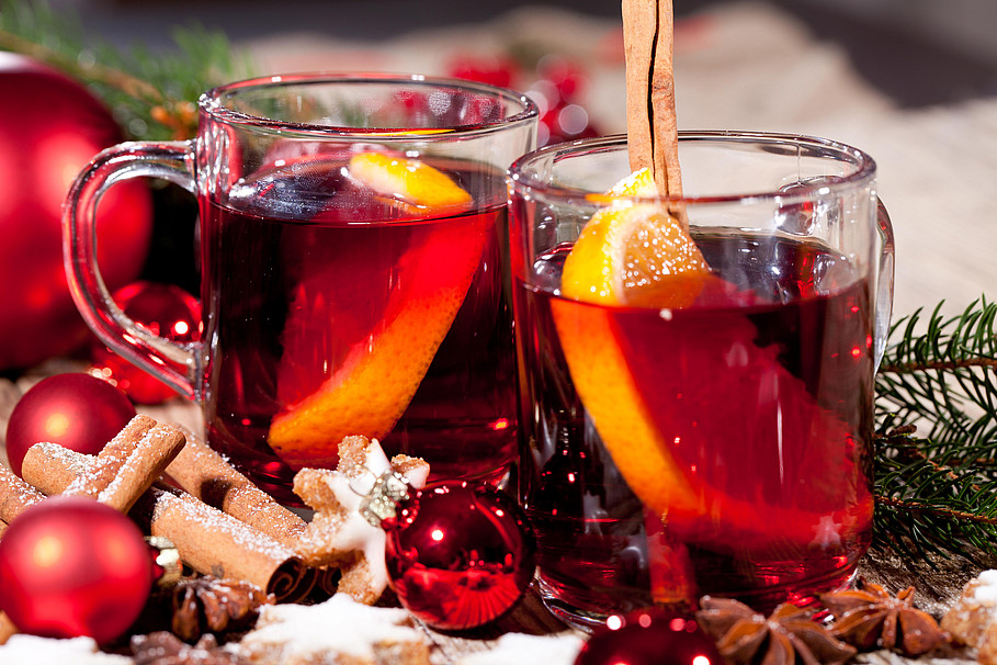 Zwei Gläser Glühwein mit Orangenscheiben und weihnachtlicher Deko im Hintergrund