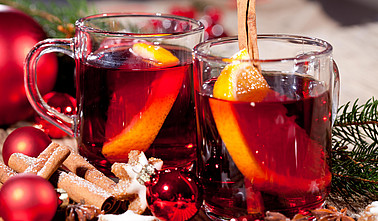 Zwei Gläser Glühwein mit Orangenscheiben und weihnachtlicher Deko im Hintergrund