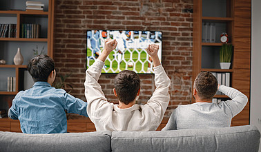 Blick von hinten auf drei Männer auf einer Couch vor dem Fernseher, wo Fußball läuft.