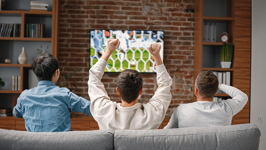 Blick von hinten auf drei Männer auf einer Couch vor dem Fernseher, wo Fußball läuft.
