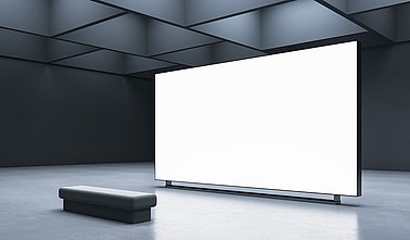 Perspektivischer Blick auf leeren, weißen, großen Bildschirm mit Platz für Werbung, Text oder Logo einer Marke in abstrakter Industriehalle mit grauer Wand im Hintergrund und Bank auf Betonboden. 