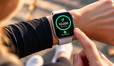 Nahaufnahme einer Smartwatch am Handgelenk, die 10.000 Schritte und das Erreichen des Tagesziels anzeigt.