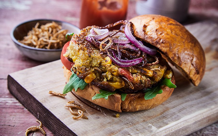 Spezialitäten-Insektenburger mit gebratenen Mehlwürmern und Salatgarnituren mit Rucola auf einem knusprig-frischen Brötchen, serviert auf einem für das Menü geeigneten Holzbrett.