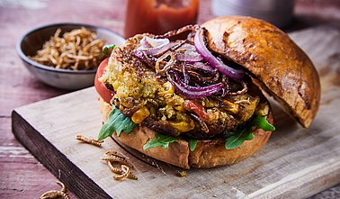 Spezialitäten-Insektenburger mit gebratenen Mehlwürmern und Salatgarnituren mit Rucola auf einem knusprig-frischen Brötchen, serviert auf einem für das Menü geeigneten Holzbrett.