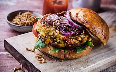 Spezialitäten-Insektenburger mit gebratenen Mehlwürmern und Salatgarnituren mit Rucola auf einem knusprig-frischen Brötchen, serviert auf einem für das Menü geeigneten Holzbrett.