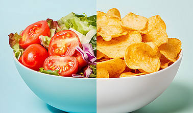 Geteilte Schale links mit frischem Salat und rechts mit Kartoffelchips auf pastellfarbenem Hintergrund. Vergleich von gesundem Essen und Junk Food.