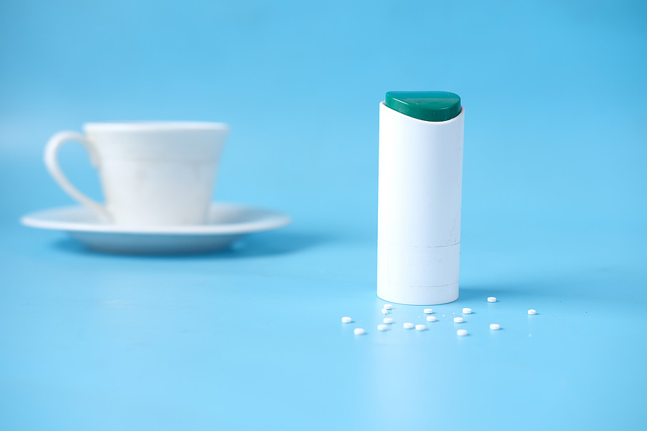 Süßstoffspender mit weißen Süßstofftabletten auf blauem Hintergrund, daneben eine unscharfe weiße Kaffeetasse mit Untertasse