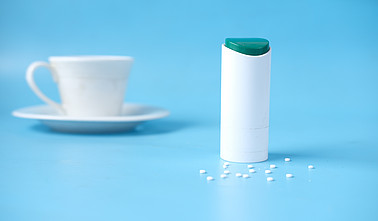 Süßstoffspender mit weißen Süßstofftabletten auf blauem Hintergrund, daneben eine unscharfe weiße Kaffeetasse mit Untertasse